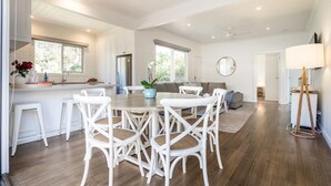 Un vaste espace de vie comprenant un coin repas avec une table entourée de chaises blanches, un canapé et une table basse. On y trouve également une cuisine équipée d’un réfrigérateur et dotée d’une fenêtre.