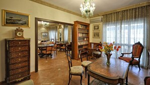 A spacious room with a large wooden dining table, multiple chairs, a chandelier, and a bookshelf filled with books.