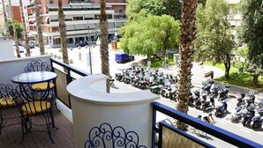 A balcony with a round table and chairs, overlooking a street with parked scooters and a few pedestrians.