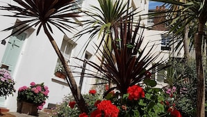 Un edificio blanco con numerosas ventanas y balcones, rodeado de plantas en macetas y flores rojas.