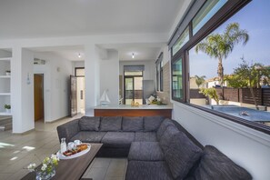 A modern living room with a large grey sectional sofa, a coffee table with food and drinks, and a view of a swimming pool through large windows. - Villa Mylos 8 is located in the absolute center of Protaras (Protaras)