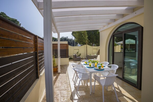 A covered patio with a table set for four, surrounded by white chairs, a wooden fence, and a glass door leading to a garden. - Villa Mylos 8 is located in the absolute center of Protaras (Protaras)
