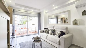 Una sala de estar moderna con un sofá, una mesa de centro y un televisor. Hay una puerta corrediza de vidrio que da a un área exterior con sillas.