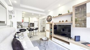 Una sala moderna con un sofá gris, un televisor y una cocina al fondo.