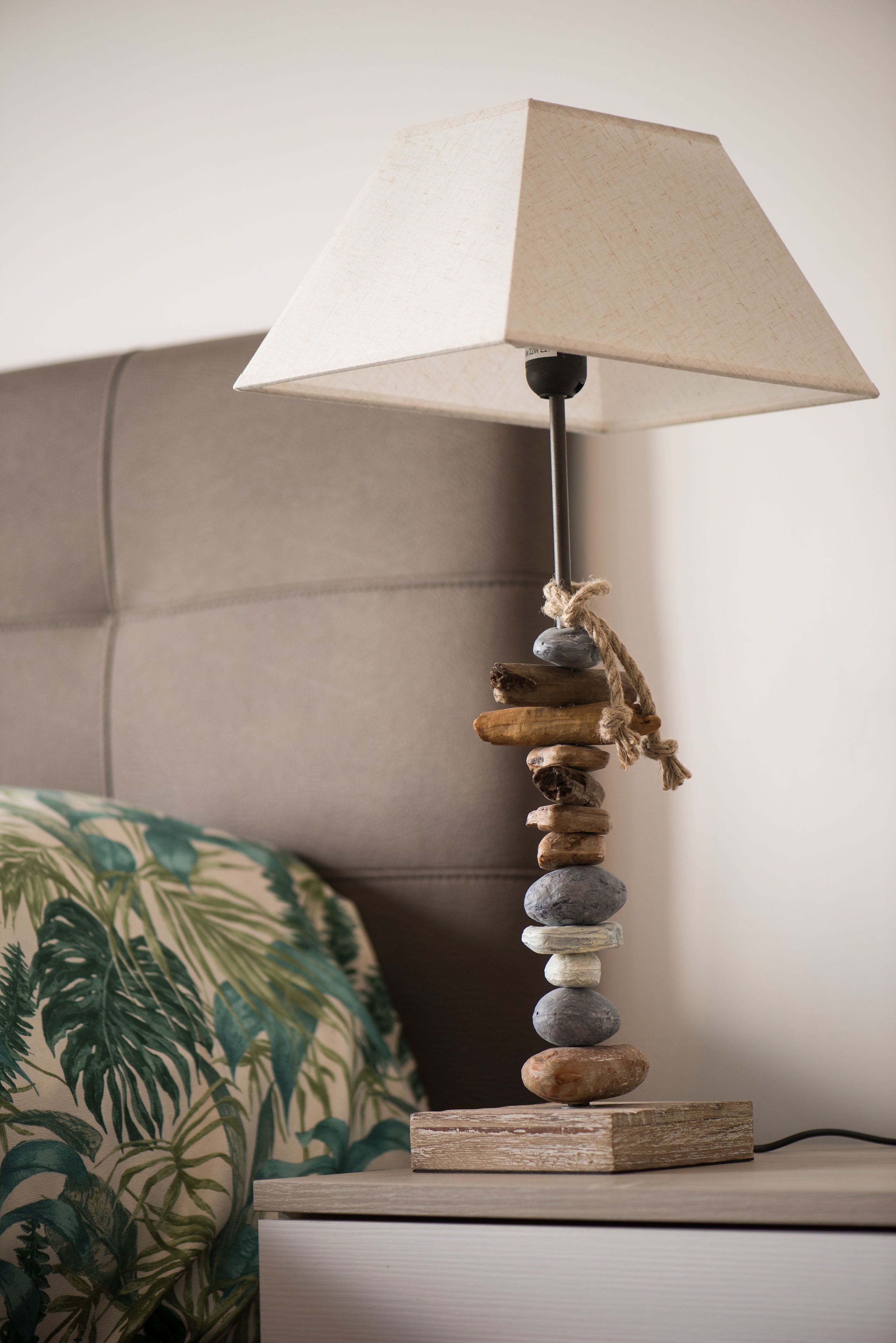 A lamp with a textured lampshade, a wooden base, and a rope tied around it, placed on a bedside table next to a bed with a floral-patterned pillow.
