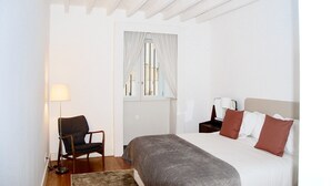 Quarto com cama, poltrona, mesa de cabeceira, abajur e janela com cortinas.