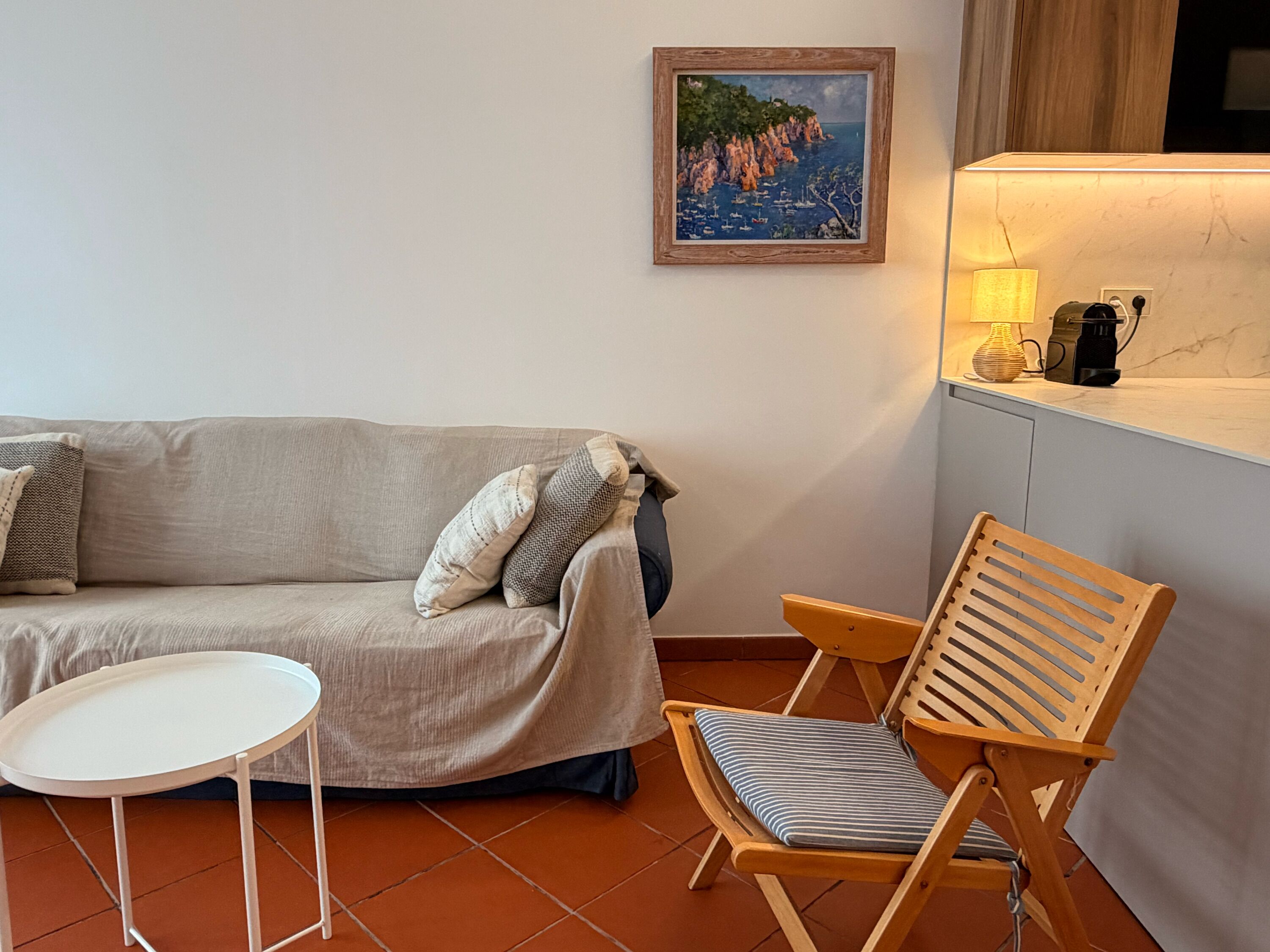 Un salón con sofá, una mesita, una silla de madera, un cuadro en la pared y una lámpara sobre una encimera.