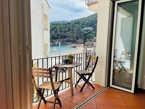 A balcony with a table and chairs, a potted plant, and a view of a beach and hillside.