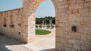 Ein Steinkorridor führt zu einem Poolbereich mit Liegestühlen und einer Pergola im Hintergrund.