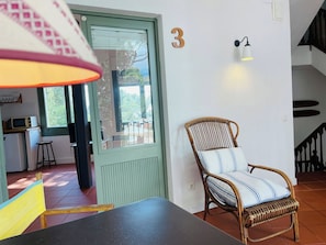 Une pièce comprenant une table à manger, une chaise en osier avec un coussin à rayures, le chiffre « 3 » peint sur le mur, un coin cuisine avec un four à micro-ondes et un escalier.