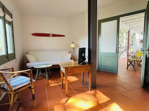 A room with a sofa, a small table, a chair, a TV, and a surfboard on the wall.