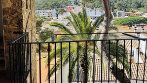Un balcón con pared de piedra, palmeras y vista a un pueblo con edificios y una colina al fondo.