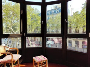 A room with a large window overlooking a city street, a wicker chair, and a small stool. - Bright 1-Bed Apartment on La Rambla, Barcelona. RJ (Barcelona)