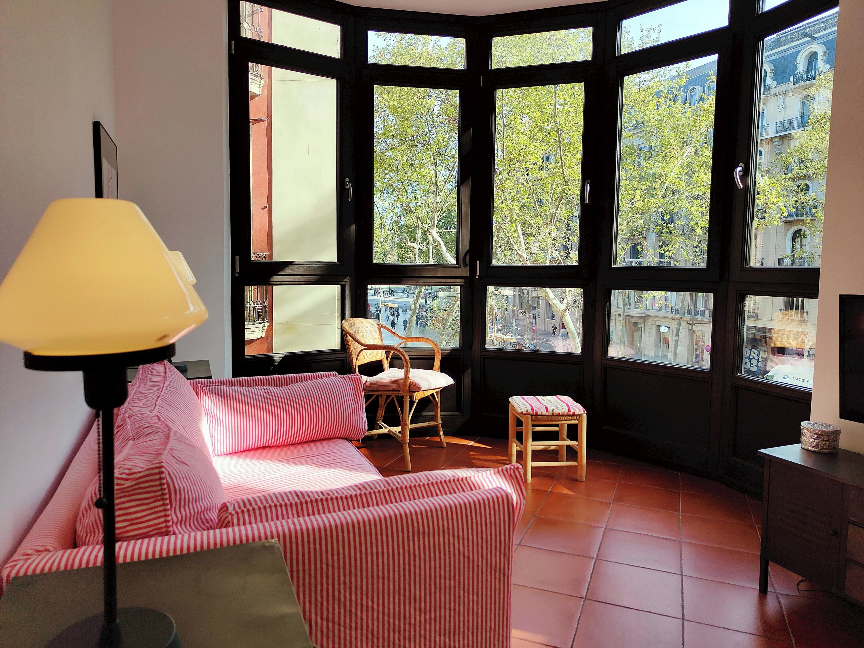 Una habitación con una cama en esquina, una silla, un taburete pequeño y una lámpara.