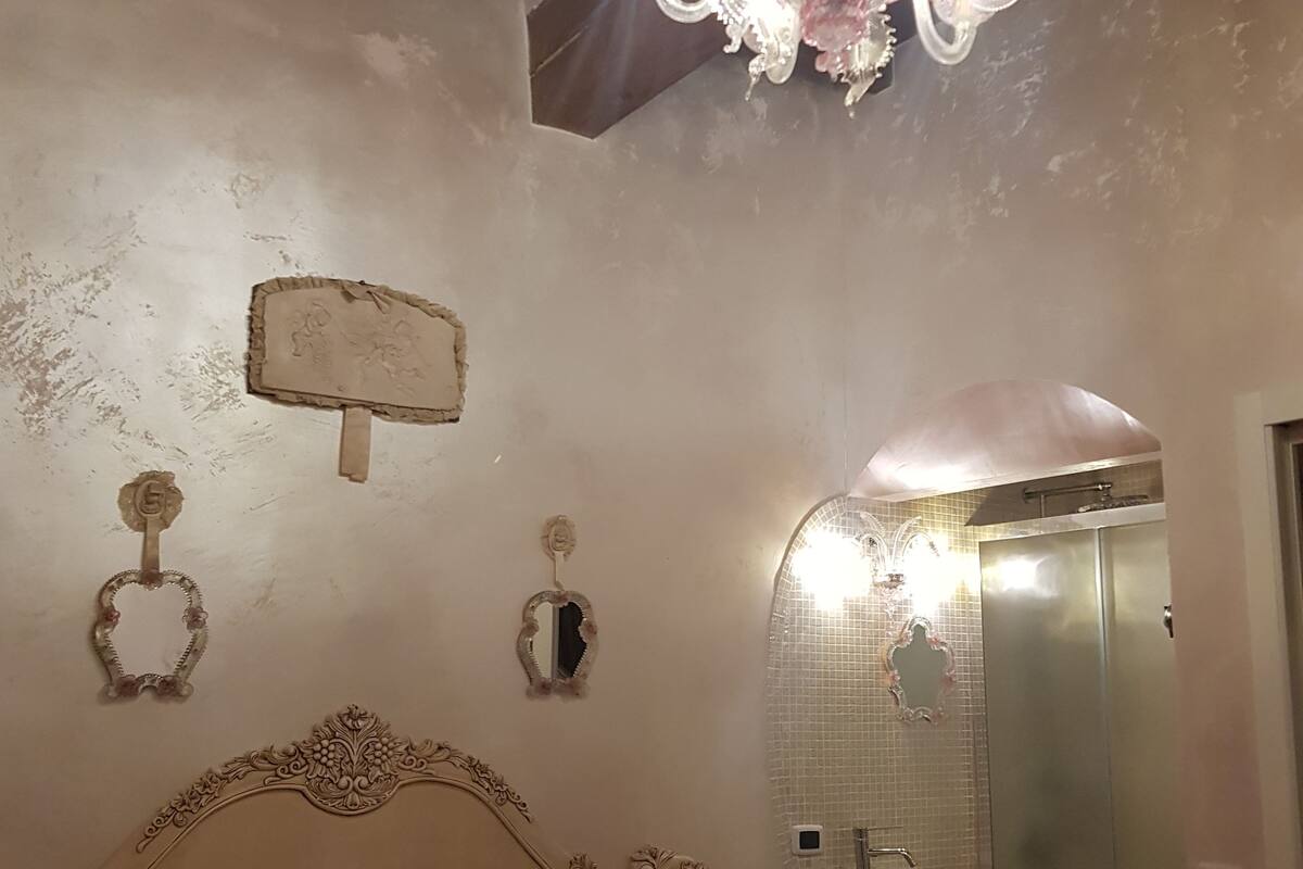 Ein Schlafzimmer mit Holzbalkendecke, einer Kristallleuchtre, einem Bett mit weißen Leinen, einem Nachttisch mit Lampe, einem an der Wand befestigten Spiegel und einem durchgehenden Türrahmen einsehbaren Badezimmer.