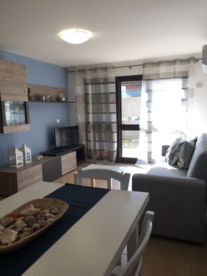 Ein modernes Wohnzimmer mit einem grauen Sofa, einem Esstisch mit weißer Tischdecke und einem Fernseher auf einer Holzkommode.