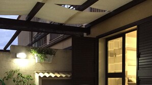 A covered patio area with a table and chairs, a potted plant, and a window with a view of the interior.