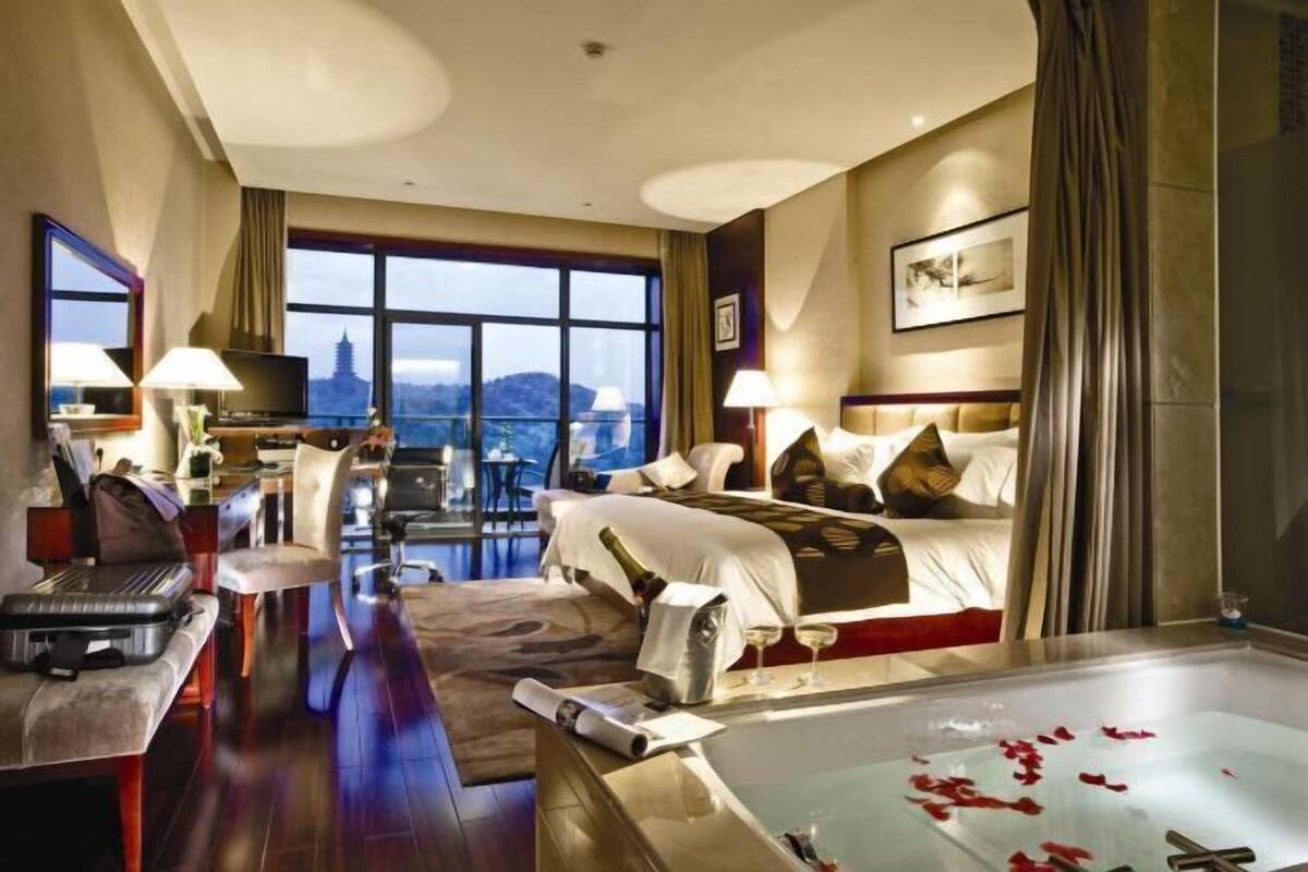 Une chambre d’hôtel avec un grand lit, un bureau avec un ordinateur, une chaise, une petite table, une baignoire remplie de pétales de rose et une vue sur une pagode, visible à travers de grandes fenêtres.