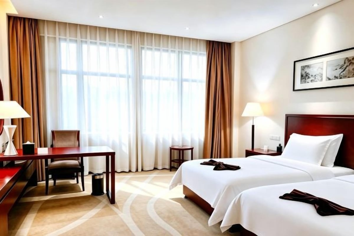Ett hotellrum med två sängar, ett skrivbord, en stol, en lampa och ett stort fönster med gardiner.