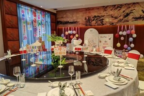 Uma mesa redonda posta com louças e copos, rodeada por cadeiras com almofadas vermelhas e brancas.