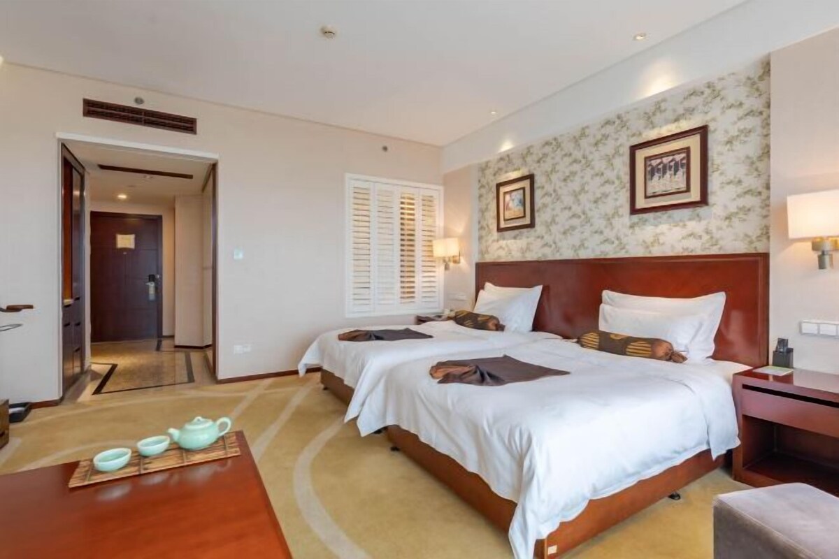 Habitación de hotel con dos camas, cabecera de madera, mesita de noche, lámpara, un cuadro en la pared y una pequeña mesa con tazas de té.