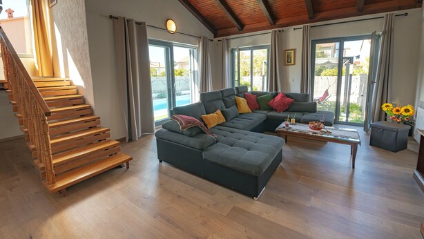 A spacious living room with a wooden staircase, a sectional sofa, and a large window offering a view of the outdoors.