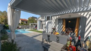 An outdoor bar area with a pool in the background, a covered seating area, and a variety of bottles on a tray.