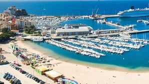 Uma marina com vários barcos atracados, uma área de praia com carros estacionados e um grande navio de cruzeiro ao fundo.