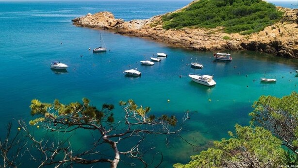 Cena costeira com barcos ancorados perto de uma ilha rochosa coberta de vegetação exuberante.