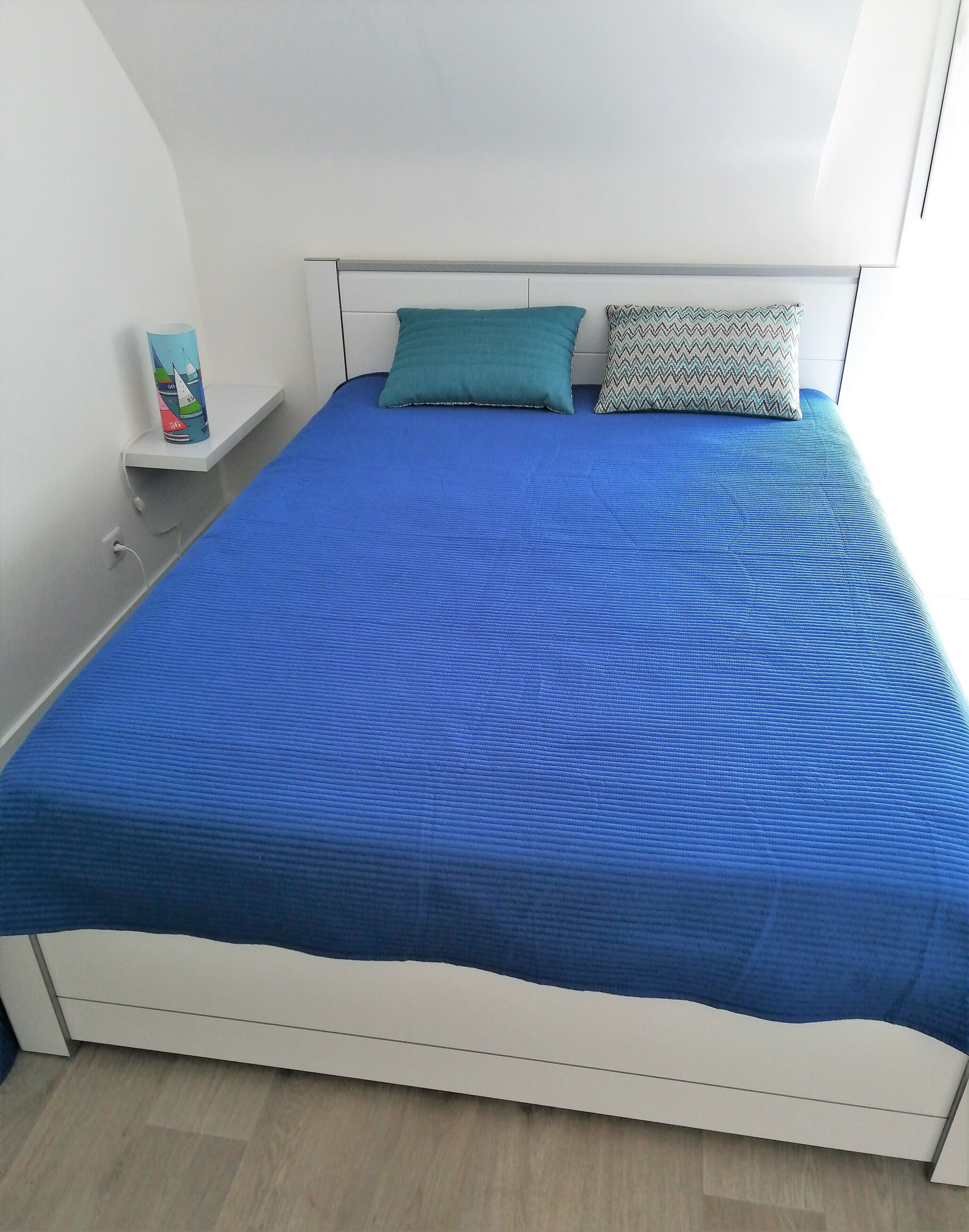 A bed with a blue bedspread and two pillows, a white nightstand with a small item on it, and a white wall.