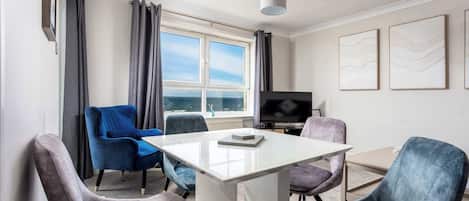 Una habitación moderna con una mesa de vidrio, cuatro sillas, un televisor de pantalla plana y un ventanal con cortinas.
