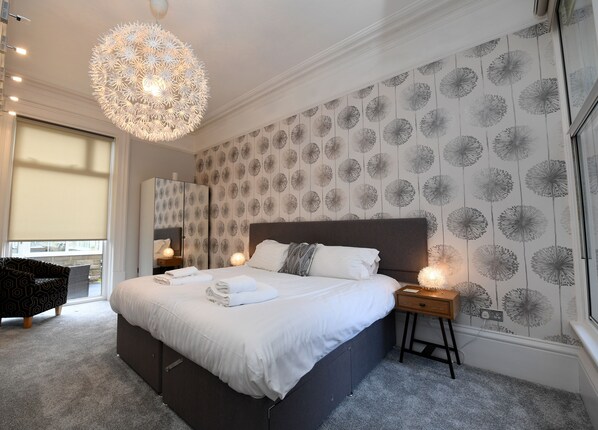 A modern bedroom with a large bed, a decorative ceiling light, a mirror, and a patterned wallpaper. - Courtyard Mews - 2 En-suite Super-king Bedrooms - Courtyard Garden - Harrogate (Harrogate)