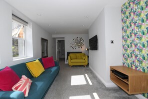 A modern living room with a blue sofa, yellow and red cushions, a yellow armchair, and a wooden TV stand. - Courtyard Mews - 2 En-suite Super-king Bedrooms - Courtyard Garden - Harrogate (Harrogate)
