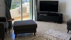 Uma sala de estar com uma televisão de tela grande, um banquinho cinza, um tapete estampado e uma porta de vidro deslizante que dá para uma varanda.