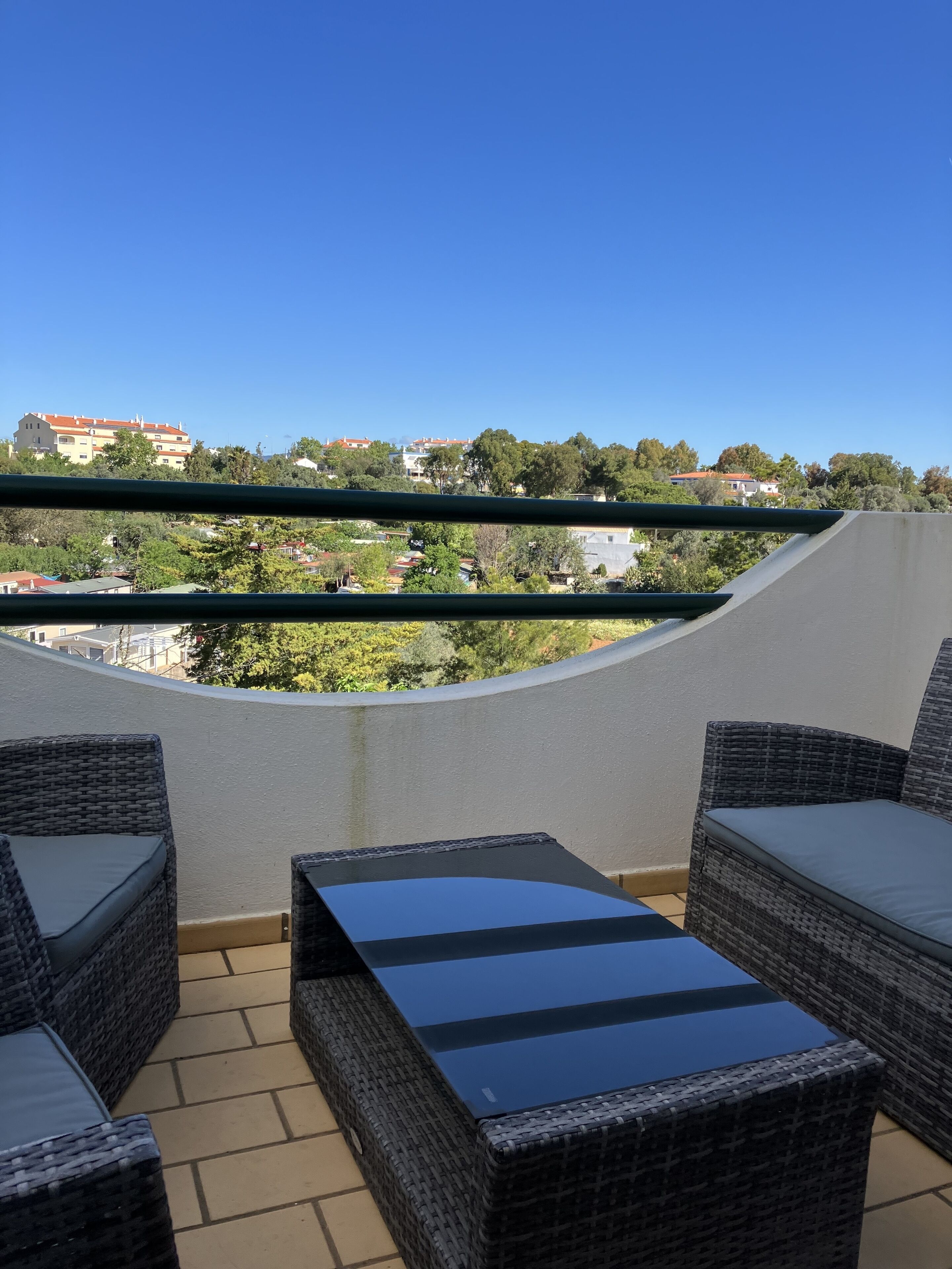 Varanda com móveis de vime e mesa de vidro, com vista para uma área residencial com casas e árvores.