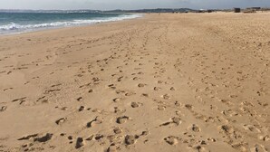Uma extensa praia de areia com pegadas, céu claro e edifícios ao longe.
