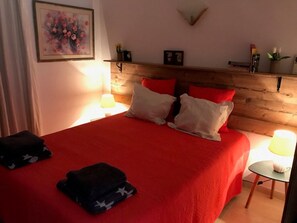 Un dormitorio con colcha roja, almohadas blancas, cabecera de madera, mesita de noche con lámpara y un cuadro en la pared.