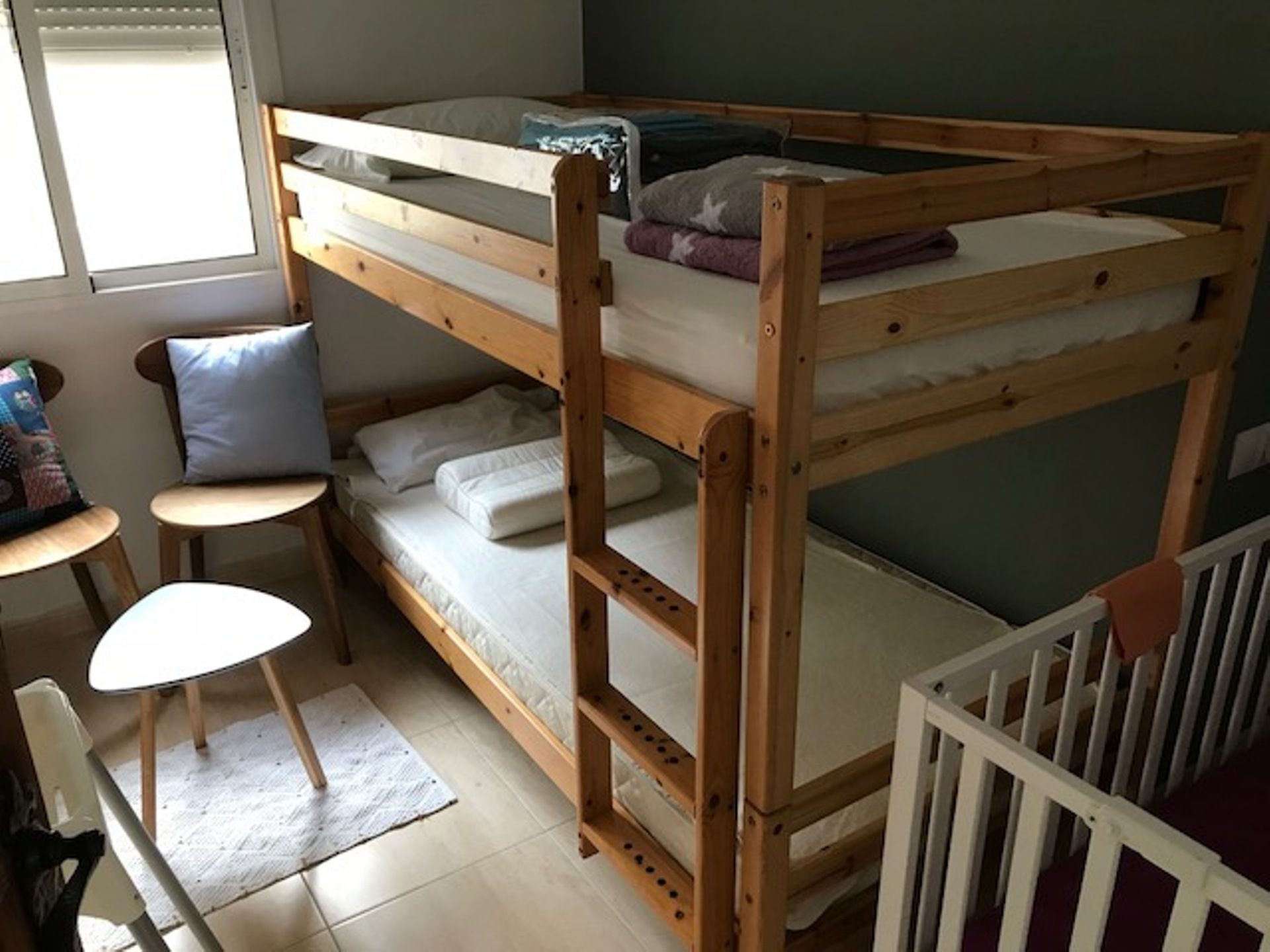 Une chambre avec un lit superposé, une petite table et une chaise, un berceau et une fenêtre.