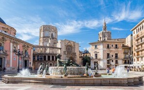 Una plaza histórica con una fuente central, una torre del reloj y arquitectura clásica circundante.