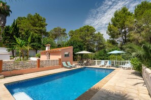 Piscina con sillas de descanso y sombrillas, rodeada de árboles y un pequeño edificio.