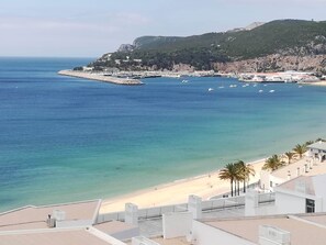 A coastal view with a beach, a rocky hill, and a pier with boats. - Apartment with terrace sea view - 2 min from the beach (Sesimbra)