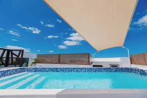 Una piscina con mosaicos azules, cerca viva de madera y un toldo grande.