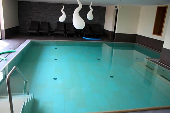 An indoor swimming pool with a clear water surface, a glass barrier, and a few lounge chairs against a dark wall. - Weinberghotel Nalbach (Reil)