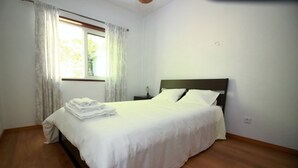 A neatly made bed with white linens, a wooden headboard, and a window with sheer curtains.