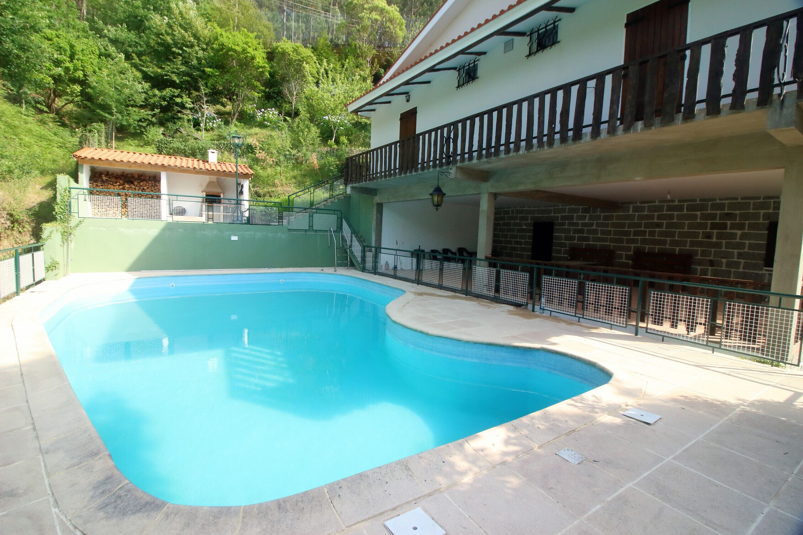 Una piscina con bordo curvo, circondata da un solarium in pietra. Si intravede sullo sfondo un edificio di due piani con un balcone e un piccolo deposito con tetto in tegole.