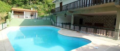 Una piscina con bordo curvo, circondata da un solarium in pietra. Si intravede sullo sfondo un edificio di due piani con un balcone e un piccolo deposito con tetto in tegole.