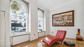 Een ruime kamer met grote ramen, een rode leren fauteuil, een groot schilderij in een lijst en een vaas op de vloer.