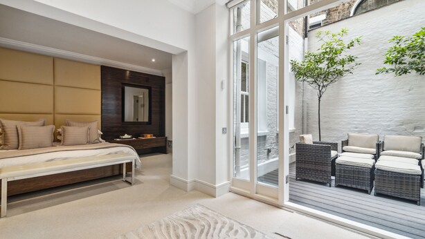 Een ruime slaapkamer met een groot bed, een bankje en uitzicht op een balkon met rieten meubels en een boom.