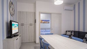 A modern kitchen with a wooden cabinet, a television, a dining table with blue chairs, and a sofa with floral cushions.