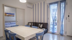 A modern room with a wooden dining table, blue chairs, a sofa with patterned cushions, and a sliding glass door leading to an outdoor area.
